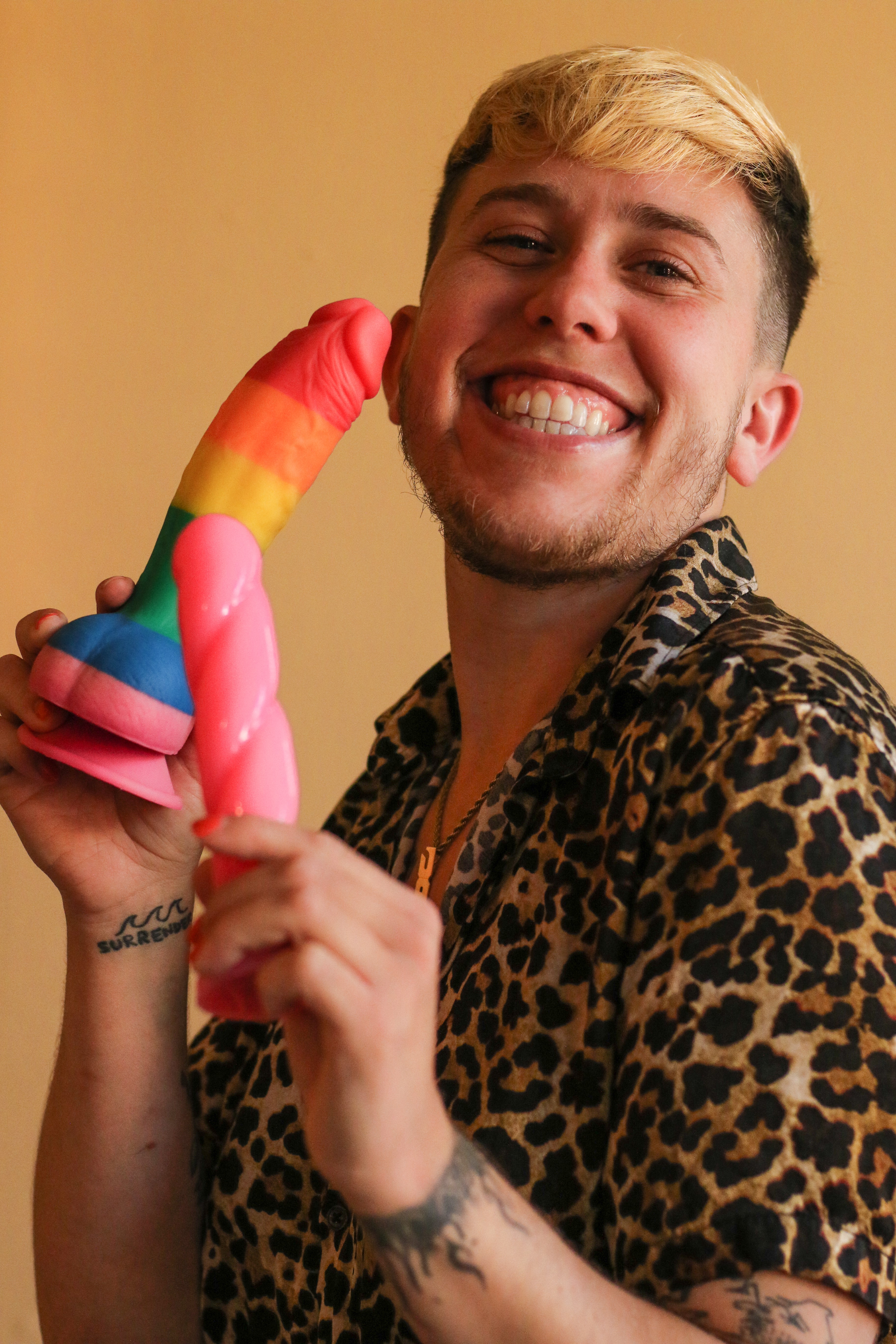 Jamie Joy (they/them) smiling and holding a rainbow dildo